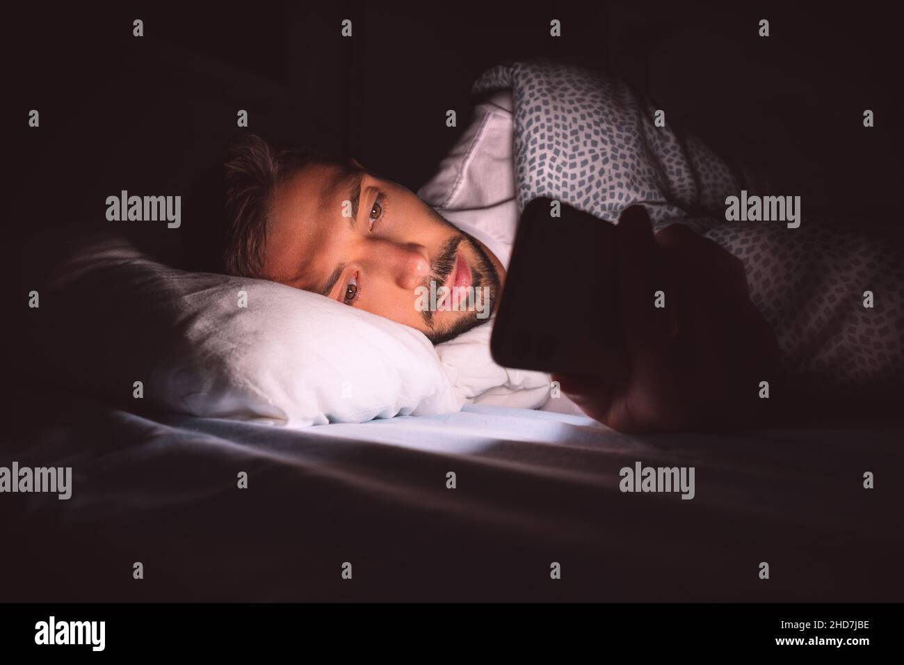 Giovane uomo sdraiato a letto e con smartphone durante la notte prima di dormire. Dipendenza dai social media e insonnia. Comunicazione e concetto di persone Foto Stock