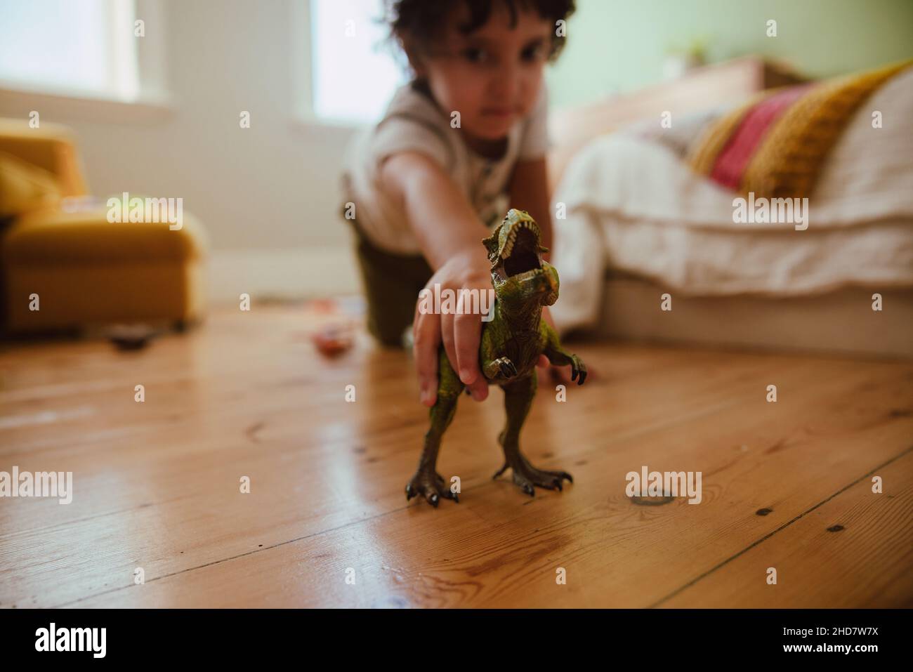 Ragazzo etnico che gioca con un dinosauro t-rex in casa. Adorabile ragazzo giovane che tiene un giocattolo ruggente di dinosauro mentre si inginocchiava nella sua area di gioco. Foto Stock
