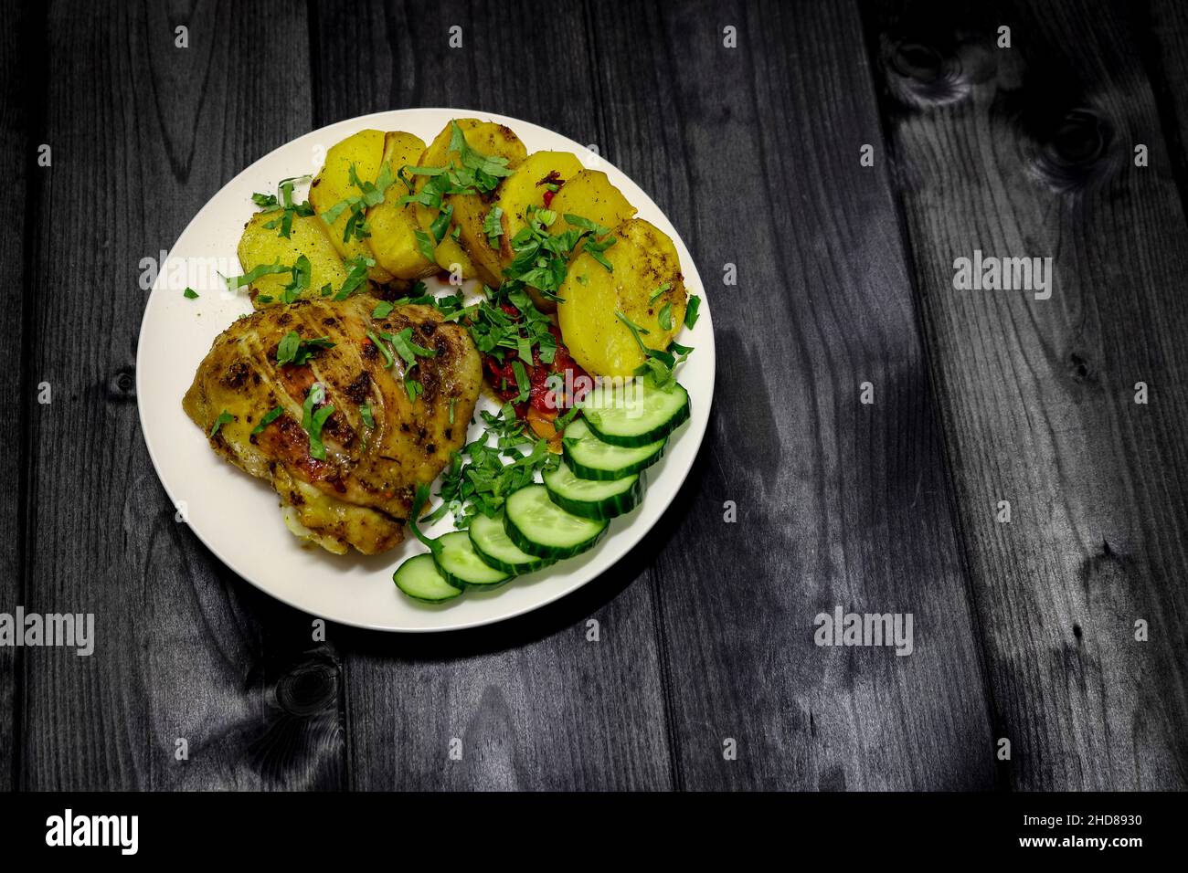 Un piatto con pollo e patate al forno, guarnito con prezzemolo verde e cetriolo a fette, vista dall'alto. Su sfondo di legno scuro. Foto Stock
