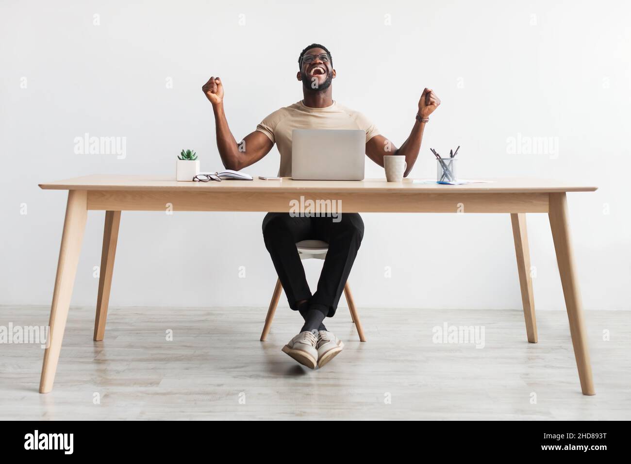 Giovane uomo nero eccitato gesturando sì di fronte al laptop, festeggiando il successo o il successo, seduto alla scrivania in ufficio Foto Stock