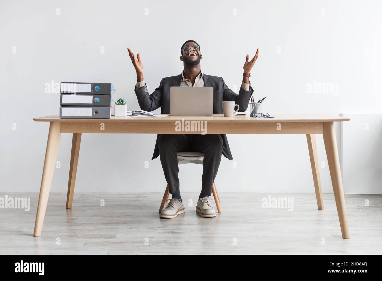 Ritratto a tutta lunghezza di un giovane e gioiato uomo d'affari afroamericano seduto al tavolo con un computer portatile contro una parete bianca Foto Stock