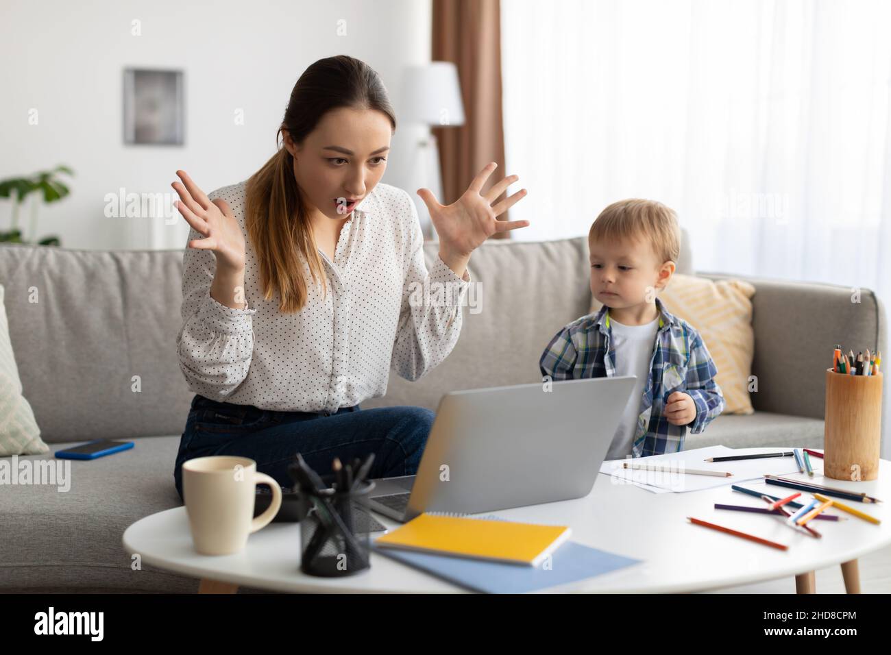 Giovane madre nervosa del toddler che lavora dalla sede, che soffre i problemi con il laptop nel paese, che reagisce emozionalmente alle notizie Foto Stock