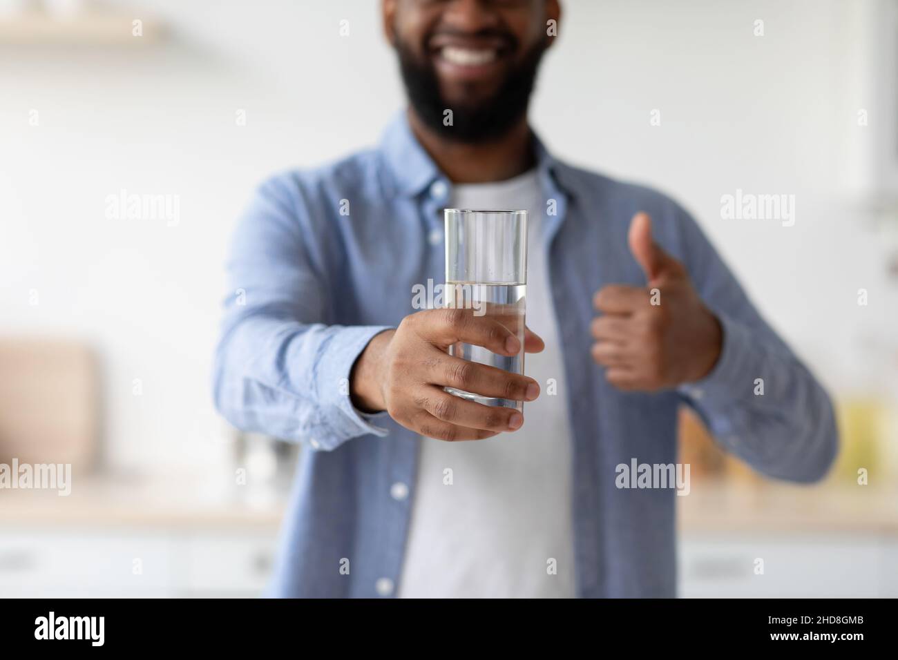 Sorridente giovane uomo nero con barba buon mattino, mostra un bicchiere di acqua pulita e pollice su Foto Stock