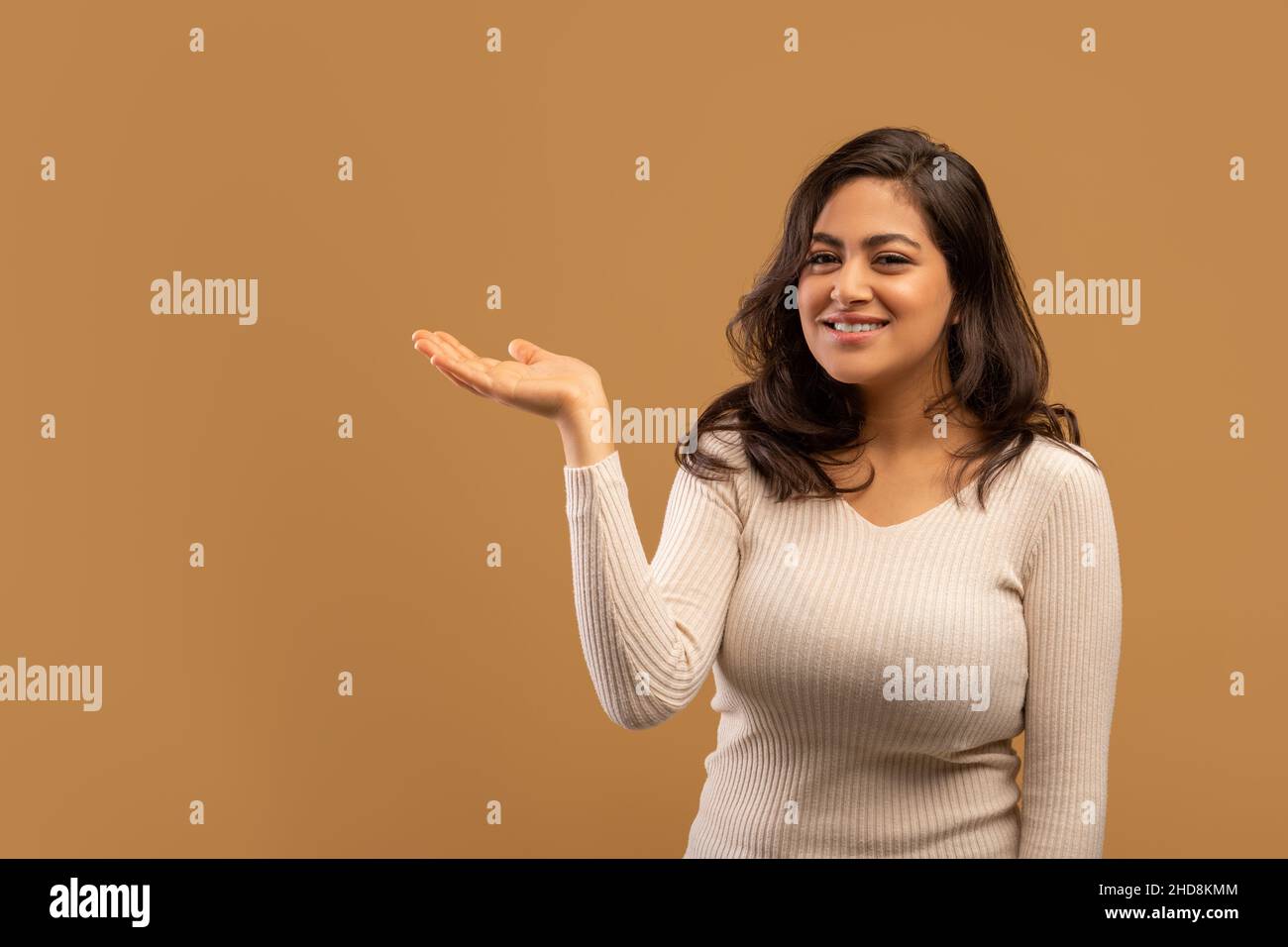 Controlla questa offerta. Positiva signora araba che tiene la sua palma aperta, dimostrando oggetto invisibile, sfondo beige, spazio libero Foto Stock