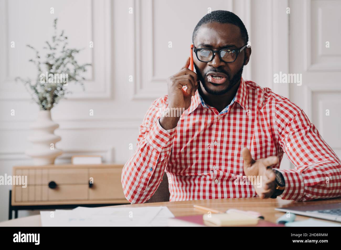 Un uomo d'affari afro-americano insoddisfatto parla emotivamente sullo smartphone mentre si siede alla scrivania Foto Stock