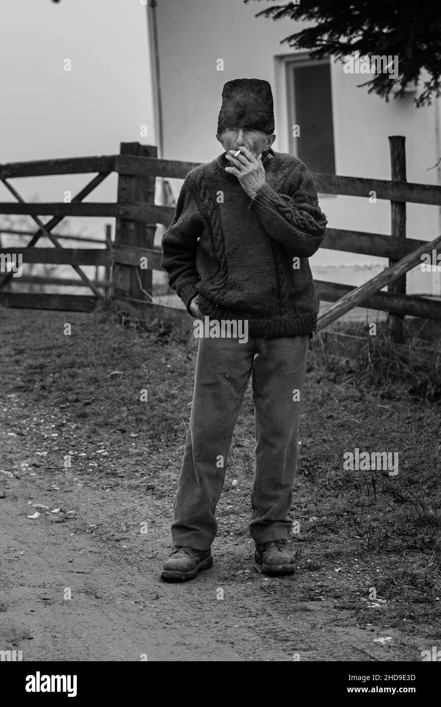 Colpo verticale di un vecchio uomo nel villaggio in piedi vicino alla strada, fumo in Romania Foto Stock