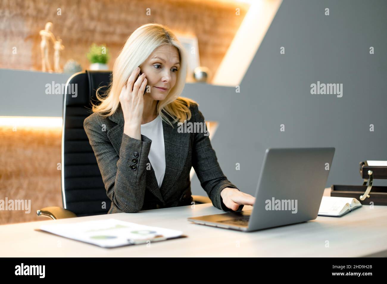 Intelligenza focalizzata sorpresa successo influente caucasica donna d'affari, freelance o top manager seduti a tavola in un ufficio utilizzando un laptop, parlando dal cellulare, navigando in internet o e-documenti Foto Stock
