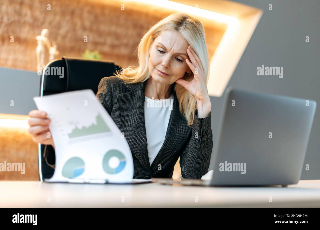 Serio successo pensivo intelligente caucasica matura donna d'affari, analista finanziario, manager, seduto alla sua scrivania in ufficio, studiando grafici finanziari, analisi e pianificazione della strategia finanziaria Foto Stock