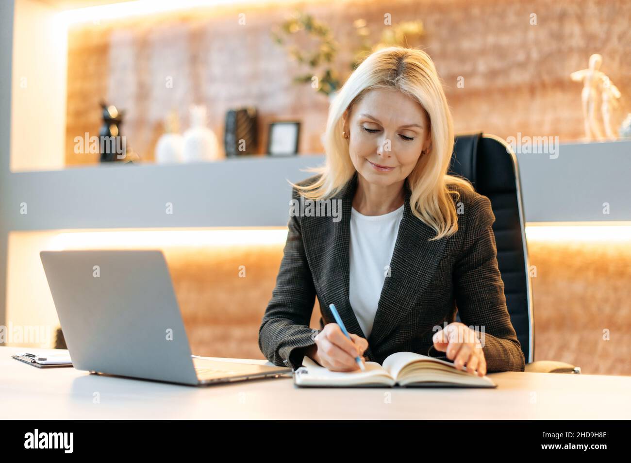 Successo intelligente attraente influente maturo caucasico bionda donna d'affari, ceo o agente immobiliare seduta in ufficio in tuta, utilizzando il laptop, prendere appunti, ponderare la strategia di business, sorridente Foto Stock