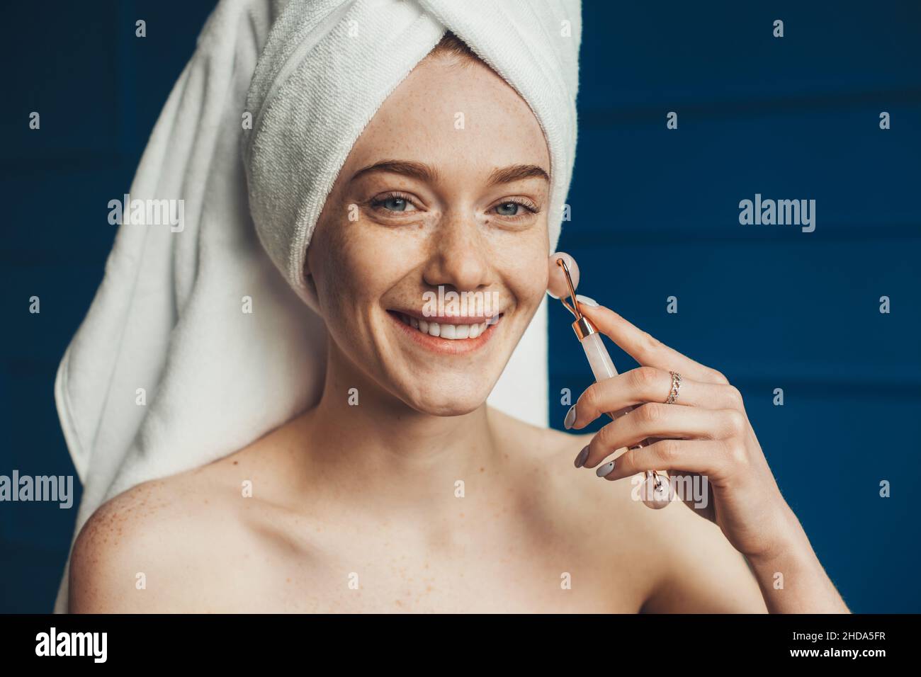 Donna caucasica ricattata avvolta in asciugamani che massaggiano il viso con rullo di giada. Primo piano verticale. Faccia sorridente. Cura della pelle. Viso di bellezza. Foto Stock