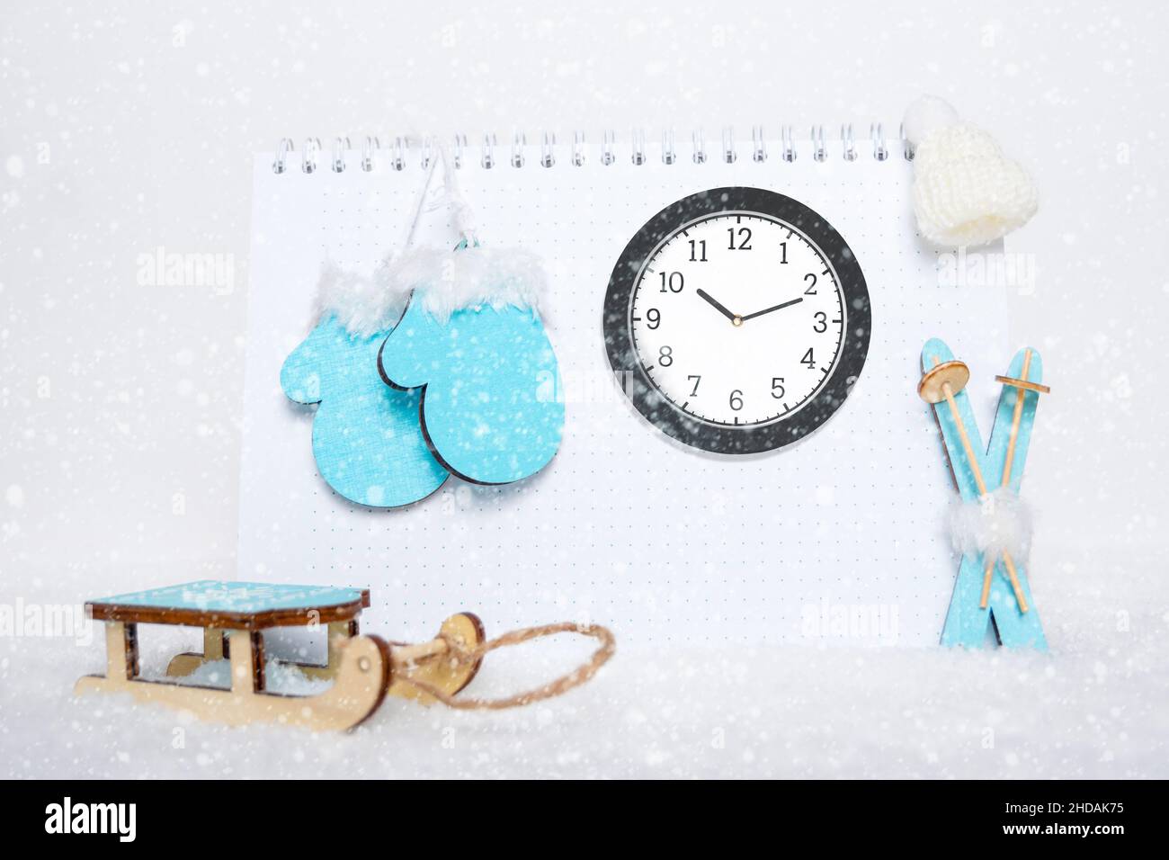 Tempo di riposo invernale. Orologio analogico rotondo, sci blu di legno giocattolo, slitte, guanti e cappello bianco su blocco note vuoto su sfondo nevoso, spazio copia. Vinello Foto Stock