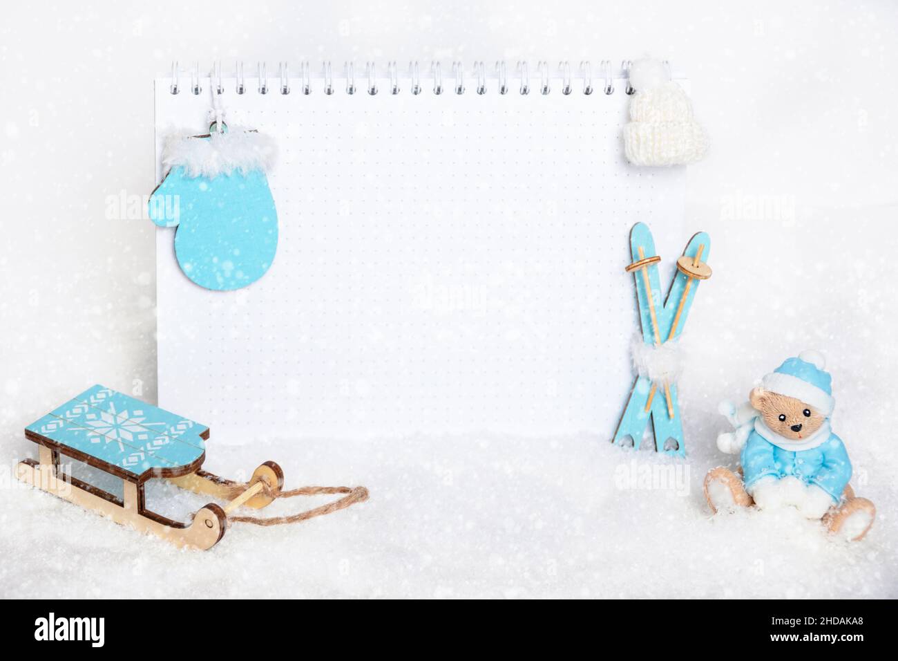 Tempo per le vacanze invernali. Taccuino vuoto, orsacchiotto che scolpisce le palle di neve, sci blu di legno giocattolo, slitte, guanti e cappello bianco su sfondo nevoso, co Foto Stock