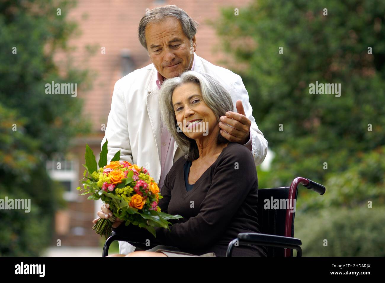 Ein älteres Paar im Grünen - die Frau sitzt im Rollstuhl und der Mann umarmt sie liebevol, Foto Stock