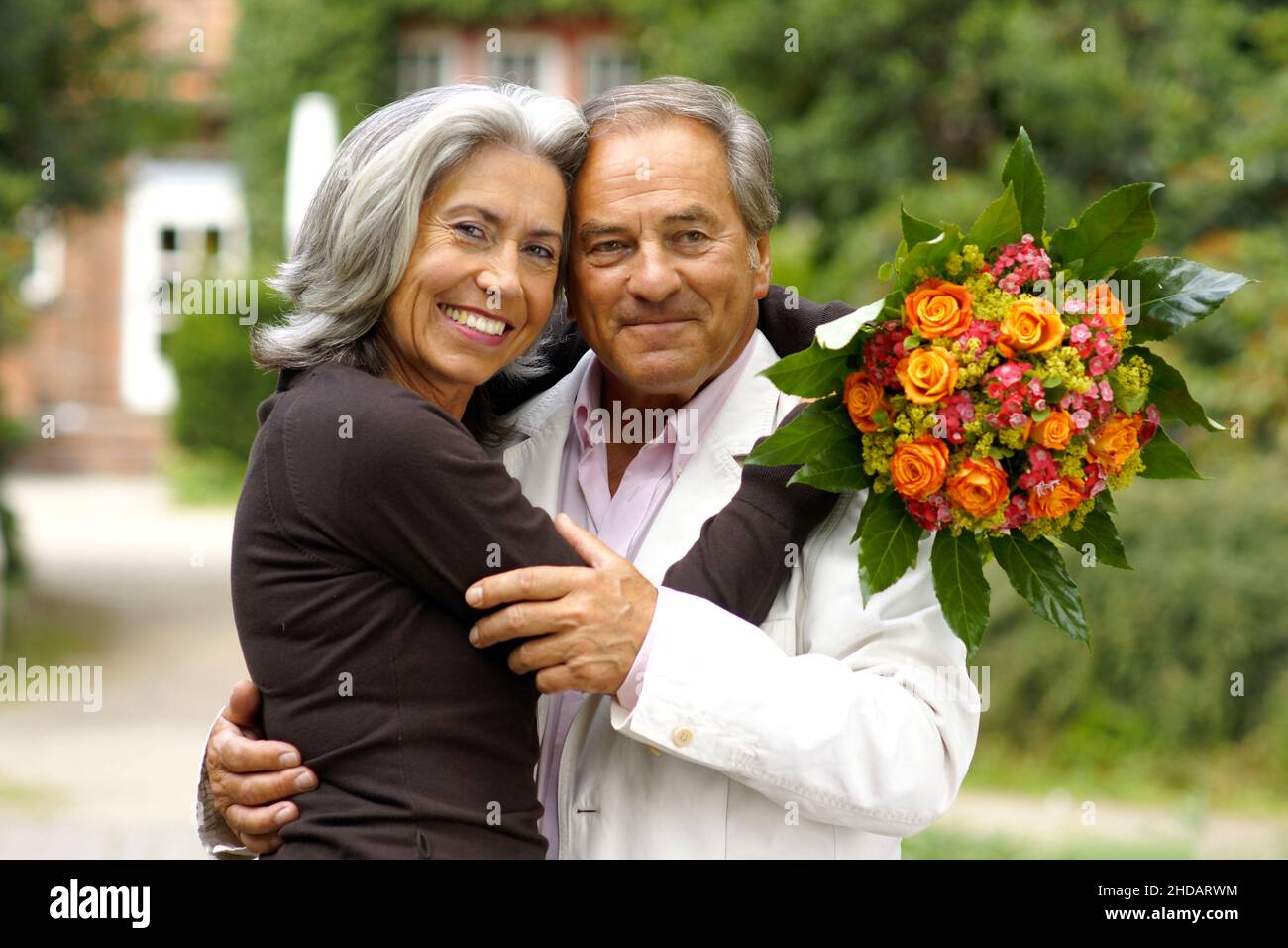 Ein aäteres Paar im Park - die Frau sitzt im Rollstuhl, der Mann umarmt sie liebevoll und ueberreicht ein Geschenk und Blumen, Model-Release| Vecchio colpo di Stato Foto Stock