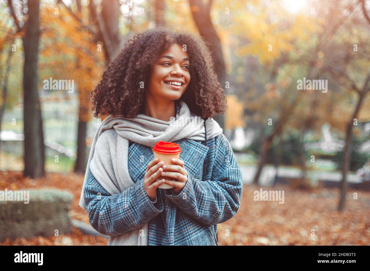 Umore autunnale. Giovane donna afro-americana felice con capelli ricci in fall Park tenendo take away caffè, guardando da parte e sorridente, rilassante in natura, Foto Stock