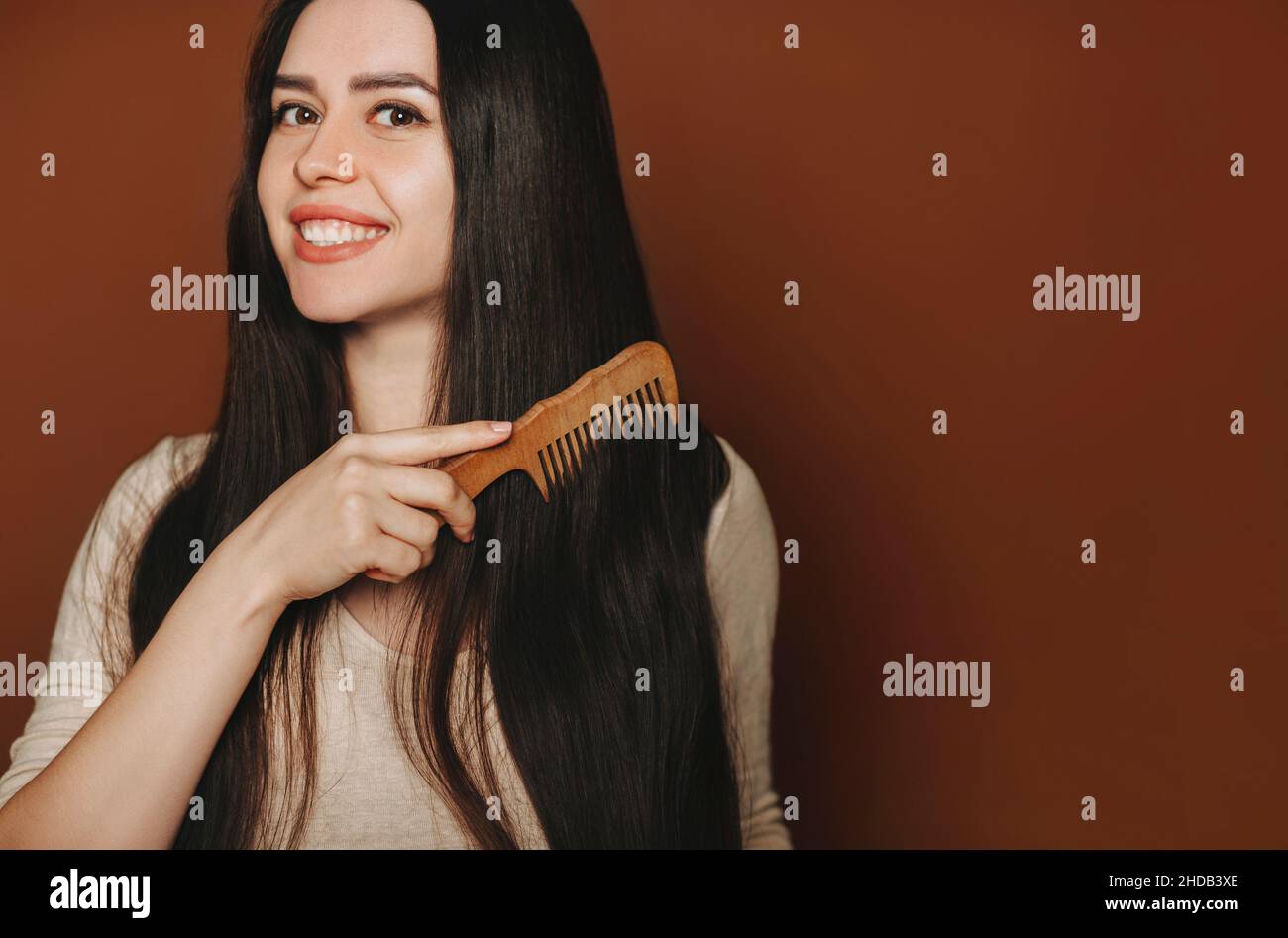 Giovane donna bruna felice pettinando i suoi lunghi capelli sani marrone e sorridendo alla macchina fotografica mentre si posa isolato su sfondo scuro con spazio copia per il testo Foto Stock