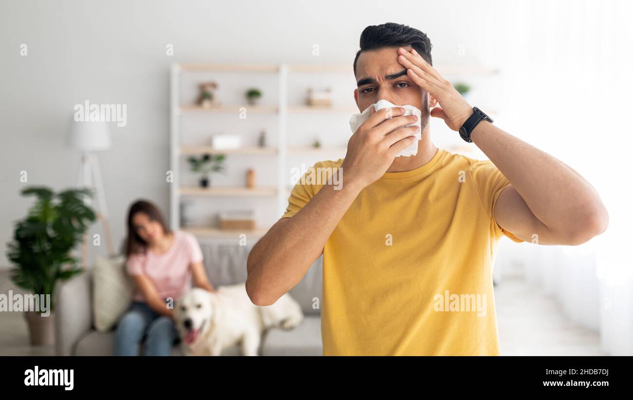 Uomo arabo che soffre di mal di testa e naso che cola, utilizzando carta tissue, avendo freddo, la sua ragazza e cane sullo sfondo Foto Stock