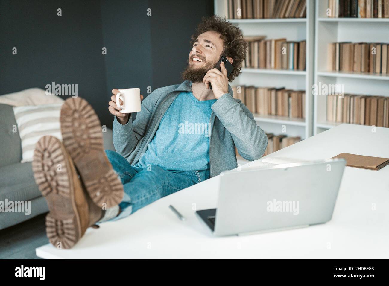 Un uomo d'affari positivo in abiti casual ha una conversazione telefonica facile con una tazza di caffè in mano e piedi sul tavolo. Solette alla videocamera. Simpatico ragazzo caucasico che parla sullo smartphone in ufficio. Primo piano. Foto di alta qualità Foto Stock
