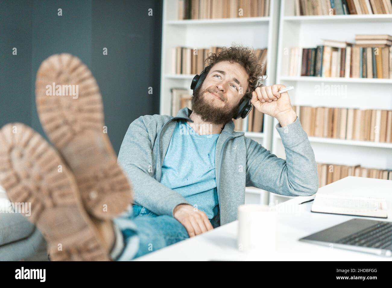 Casual Guy ascolta musica rilassante sulle cuffie nel suo ufficio, piedi sul tavolo, Soles alla fotocamera. Scaffali sfondo. Primo piano. Foto di alta qualità Foto Stock