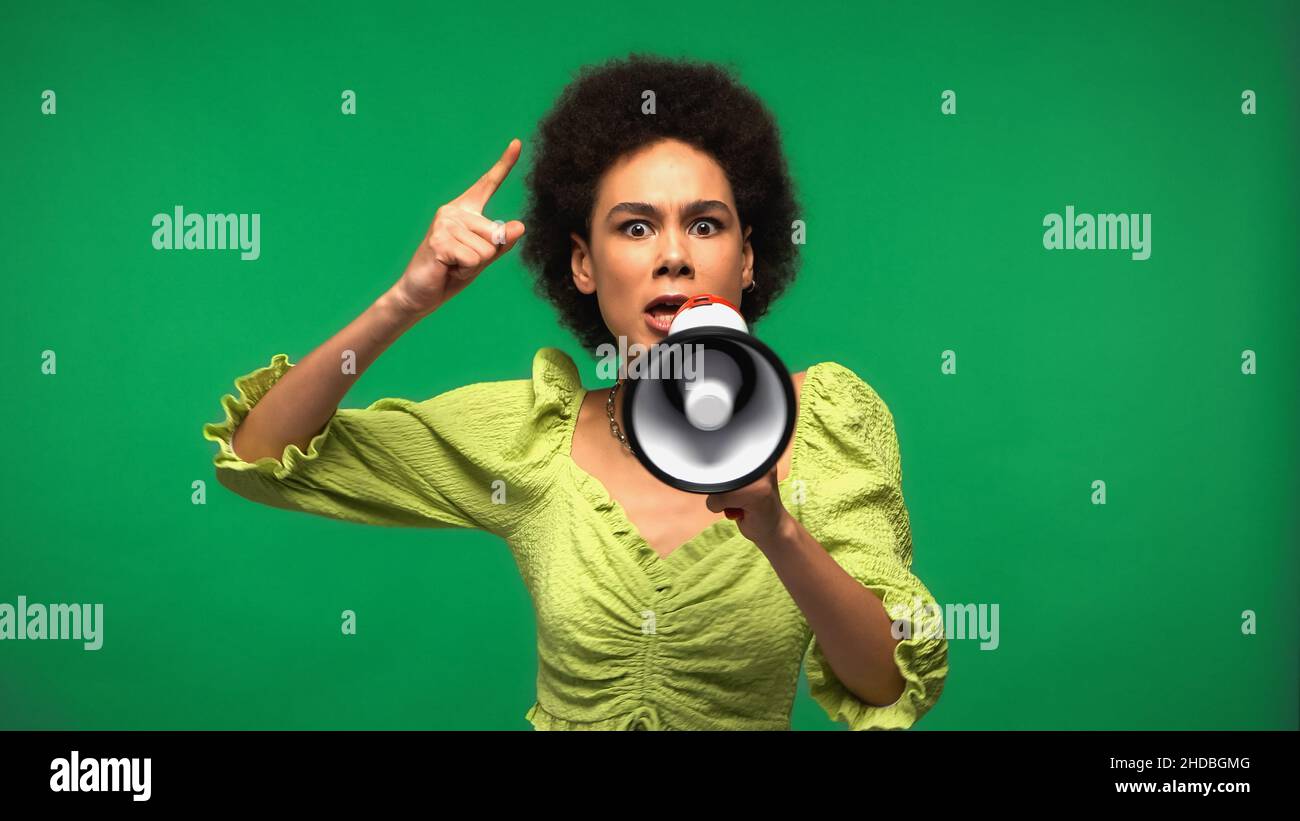 donna afroamericana che tiene il megafono e punta mentre dito mentre avvertimento isolato sul verde Foto Stock