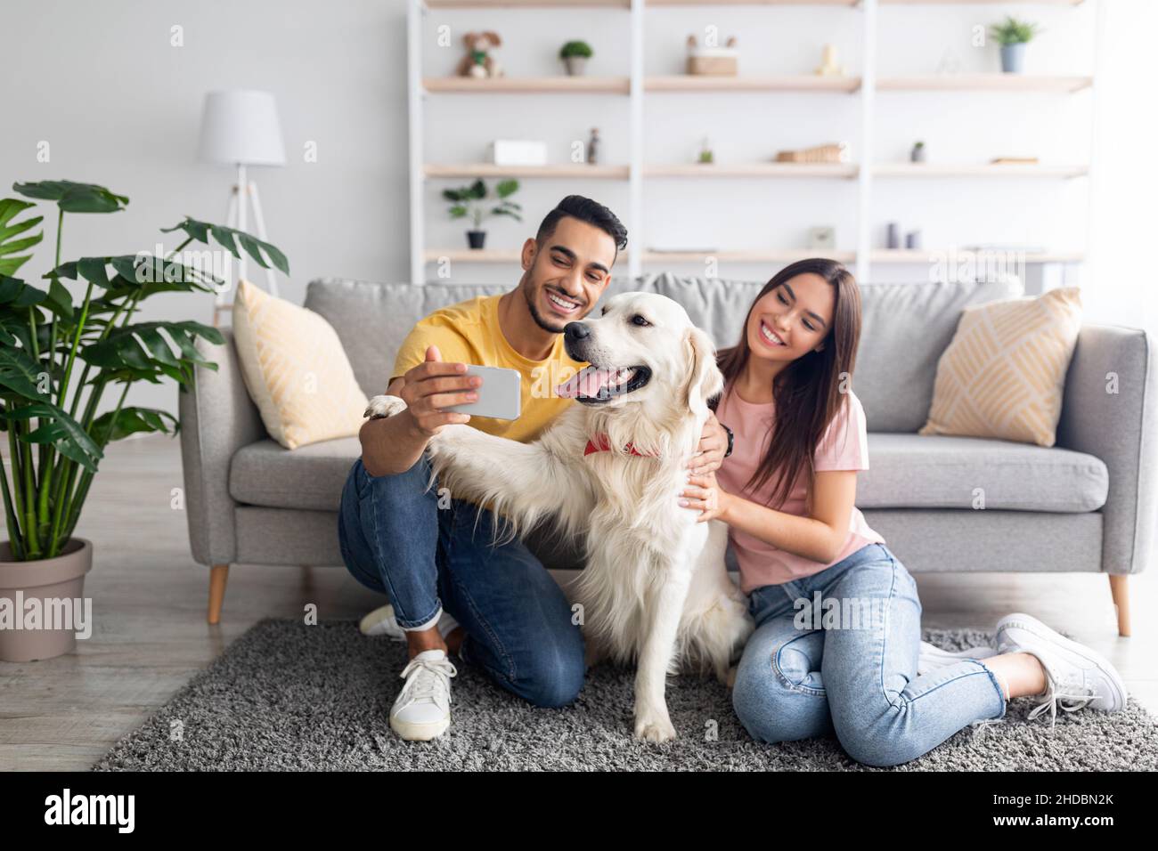 Giovane coppia internazionale che porta selfie mobili con cane da compagnia su smartphone a casa, spazio copia Foto Stock