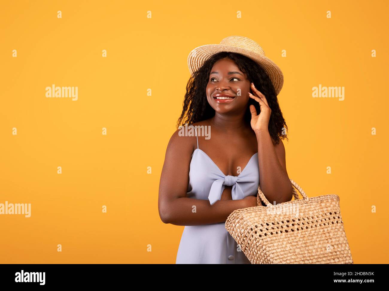 Grande offerta estiva. Bella donna nera in abito e cappello che tiene sacchetto di paglia, guardando da parte lo spazio copia su sfondo arancione Foto Stock