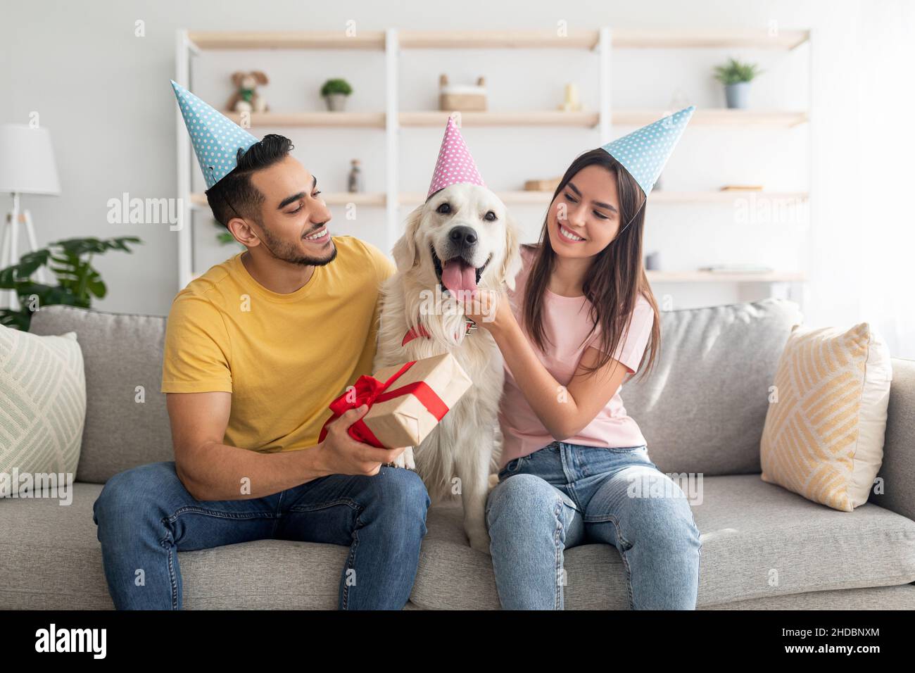 Coppia allegra interracial che celebra il compleanno del loro cane dell'animale domestico, seduta sul divano con il regalo, portando i cappelli del partito nel paese Foto Stock