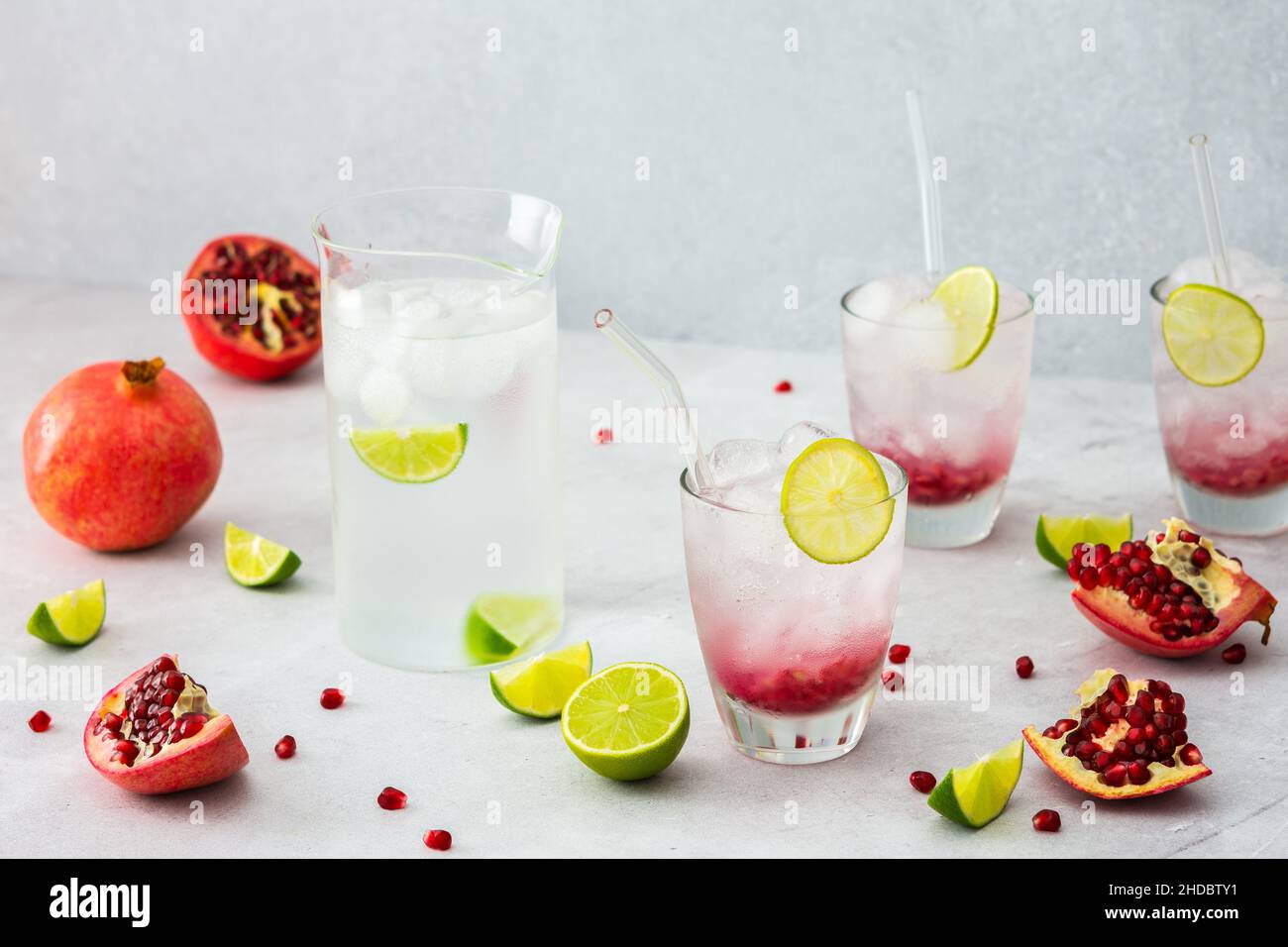 Rinfrescante bevanda di melograno in bicchieri con semi di melograno e ghiaccio, cannucce di calce e vetro, brocca di acqua fredda con agrumi, melograni tagliati Foto Stock