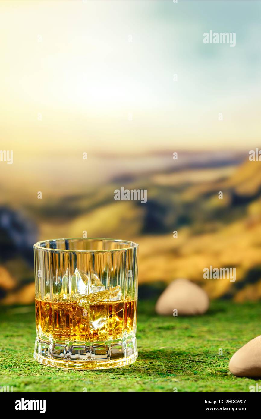 Vetro sfaccettato di whiskey affumicato posto su muschio verde con paesaggio di montagna defocused sullo sfondo Foto Stock