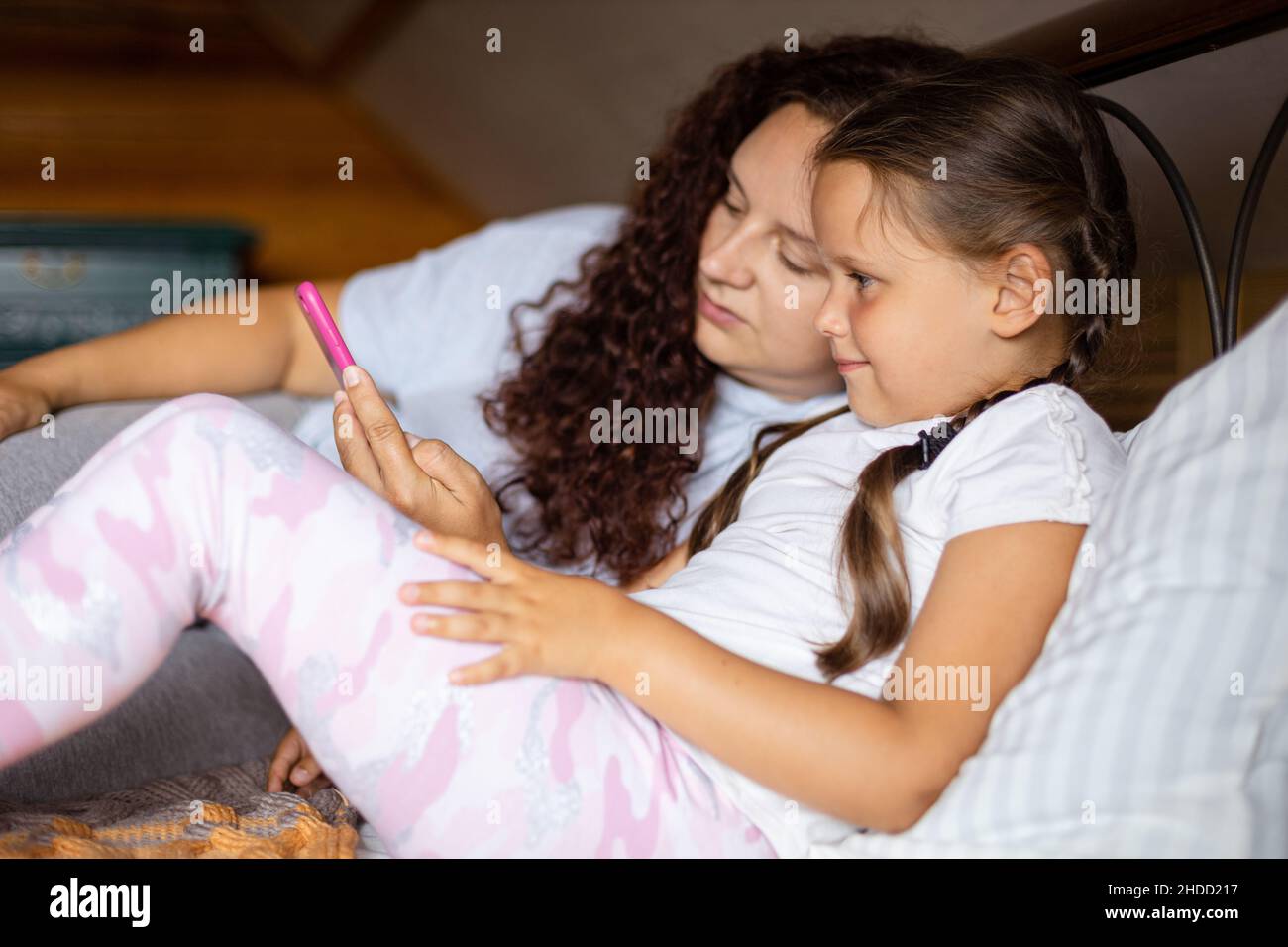 Sorridendo ragazza giovane e madre che tiene il telefono nella mano sinistra che si trova sul letto entrambi guardando il telefono in casa di legno che indossa pigiami durante il giorno. Tempo per la famiglia Foto Stock