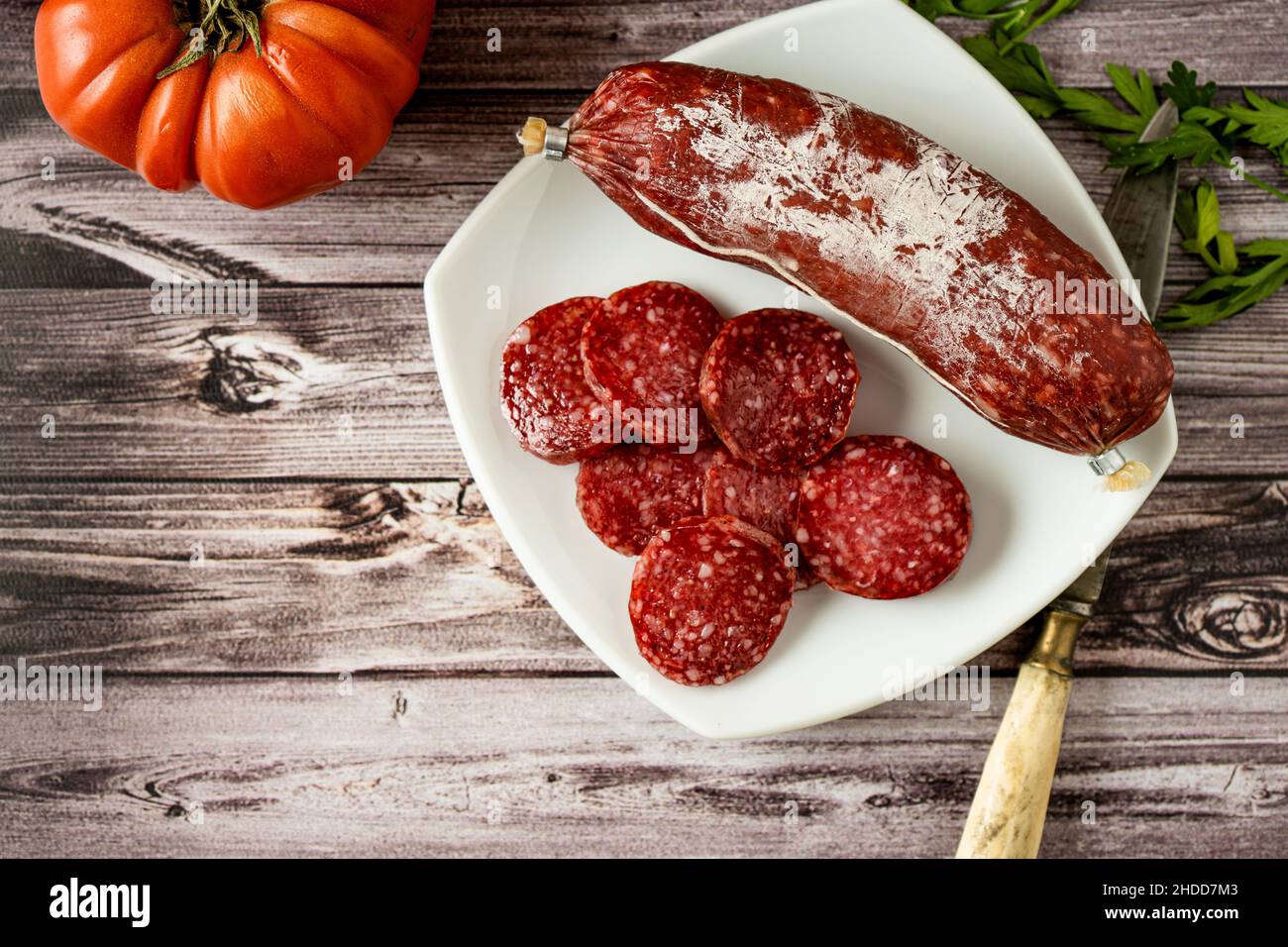 Vista aerea di Salami intero e affettato sottile su un piatto bianco su un rustico tavolo in legno. Rustico concetto di cibo di paese. Foto Stock