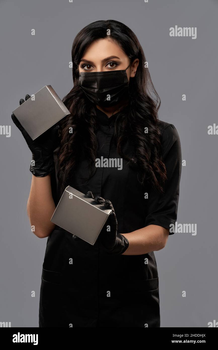 Cosmetologo professionista in uniforme nera e guanti che tengono le scatole cosmetiche in due mani mentre si trova in studio. Isolato su sfondo grigio. Foto Stock