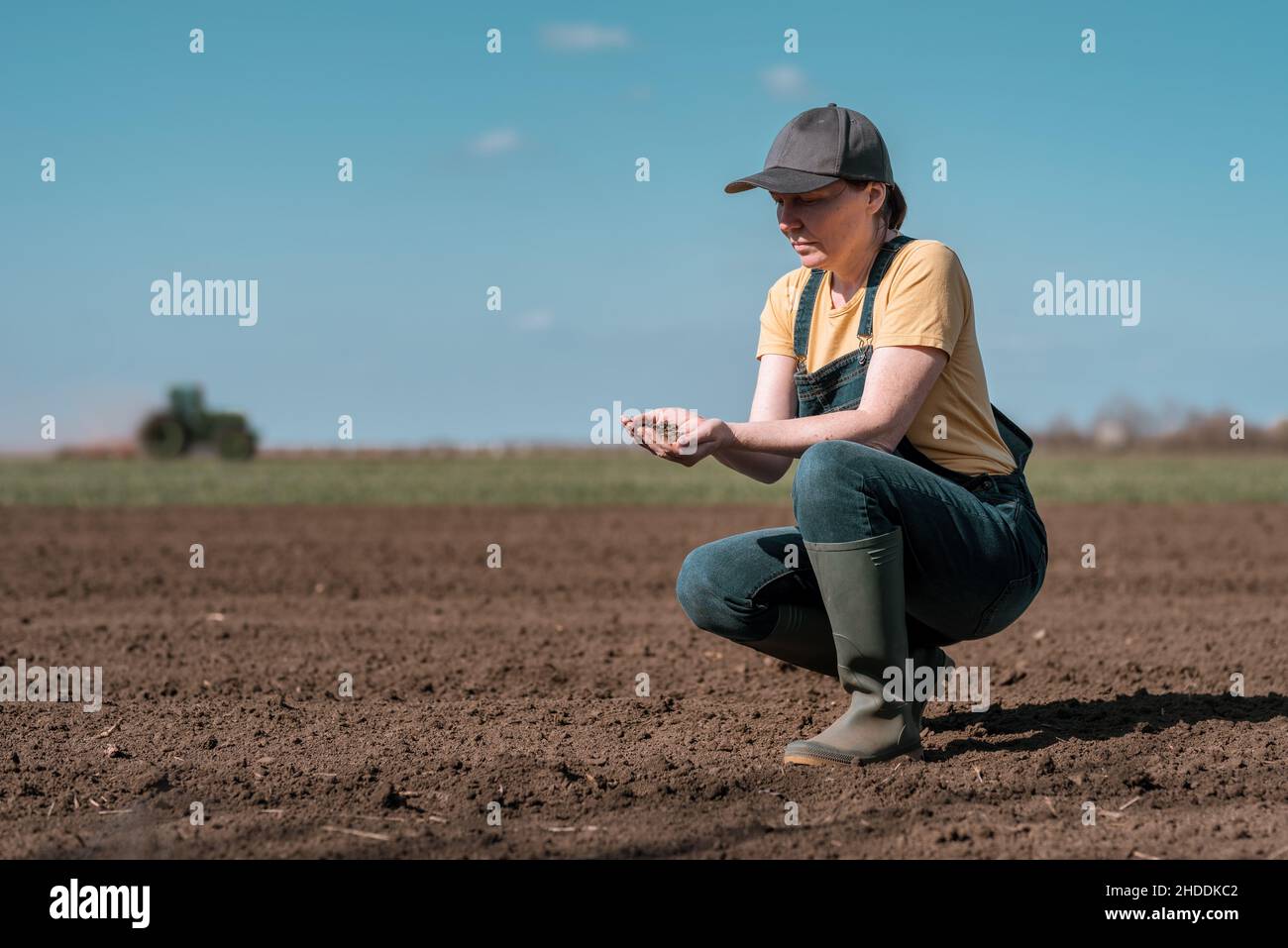 Agronomo agricoltore donna che controlla la qualità del terreno arato prima della stagione di semina, trattore agricolo in background, fuoco selettivo Foto Stock