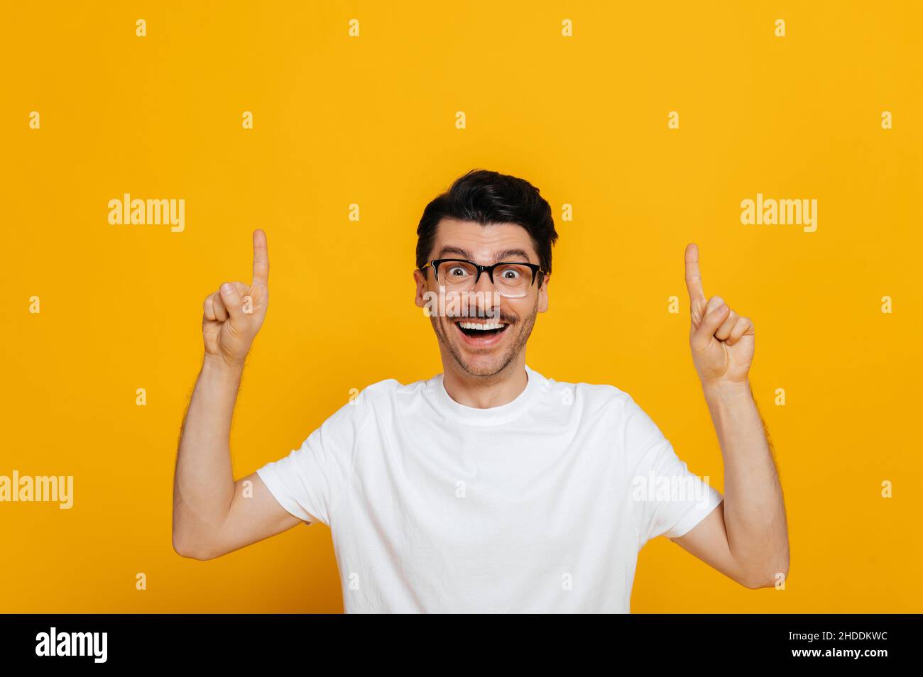 Il giovane caucasico positivo in bianco Basic t-shirt e occhiali stupiti guarda la fotocamera e punta le dita verso l'alto in uno spazio vuoto, si erge su isolato sfondo arancione Foto Stock