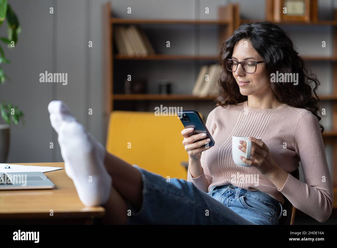 Giovane dipendente femminile rilassato godendo di una pausa di lavoro, utilizzando smartphone e bere tè sul posto di lavoro Foto Stock