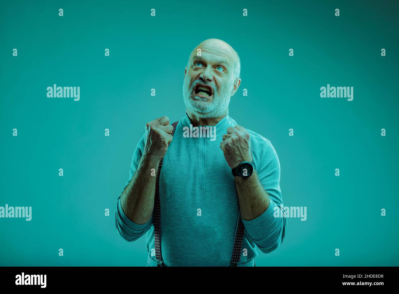 Arrabbiato frustrato uomo anziano ballando i suoi pugni e gnando i suoi denti come lui guarda su uno sfondo blu Foto Stock