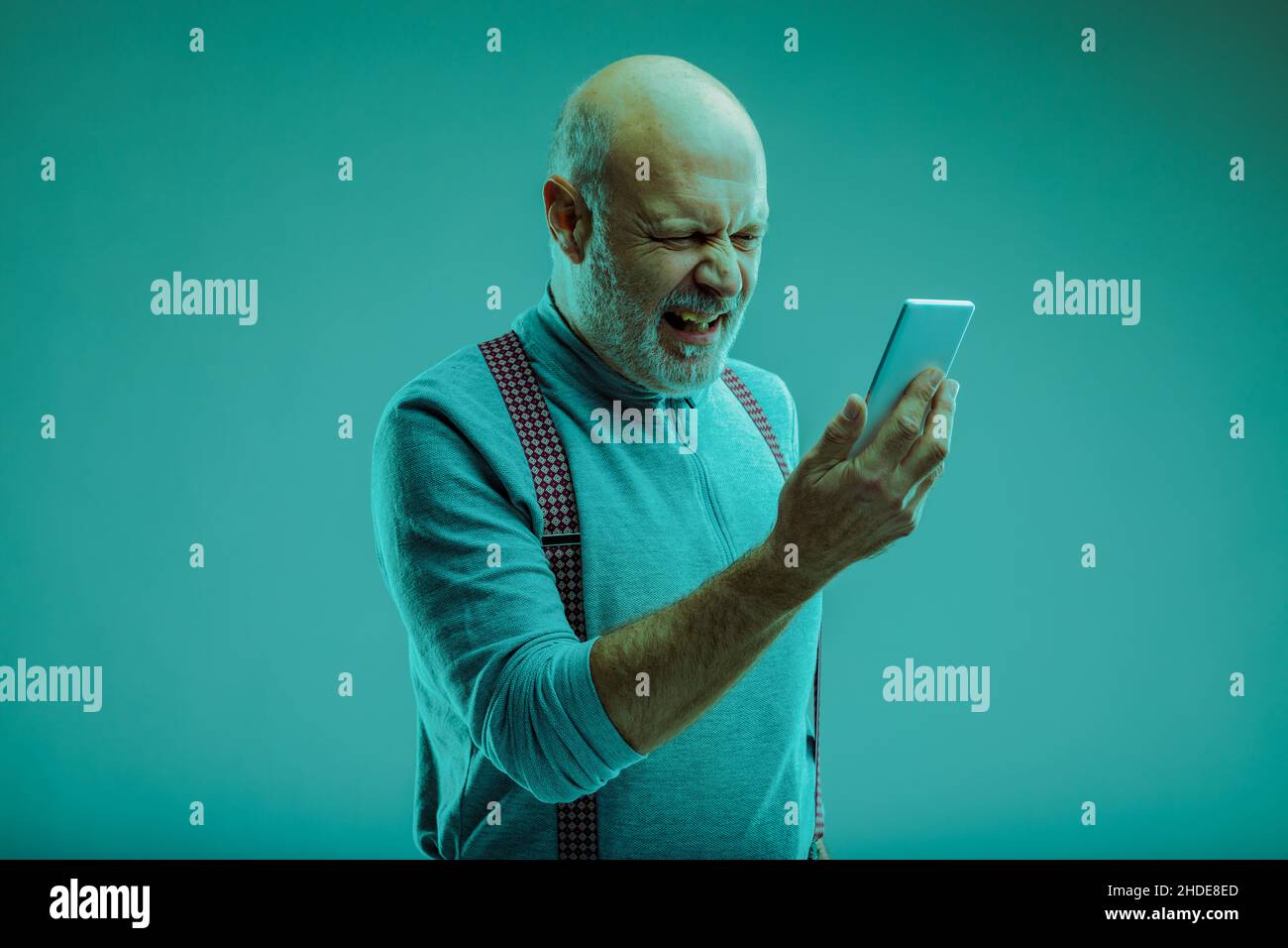 Uomo anziano che si lamenta guardando il suo smartphone in disgusto, mentre lo si accosta davanti a lui su uno sfondo blu studio Foto Stock