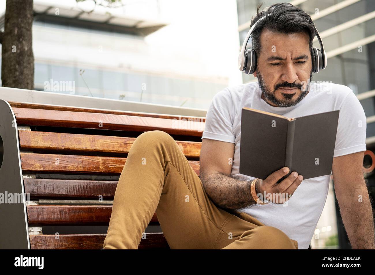 uomo latino con barba seduta su una panchina leggendo un libro in un ambiente urbano Foto Stock