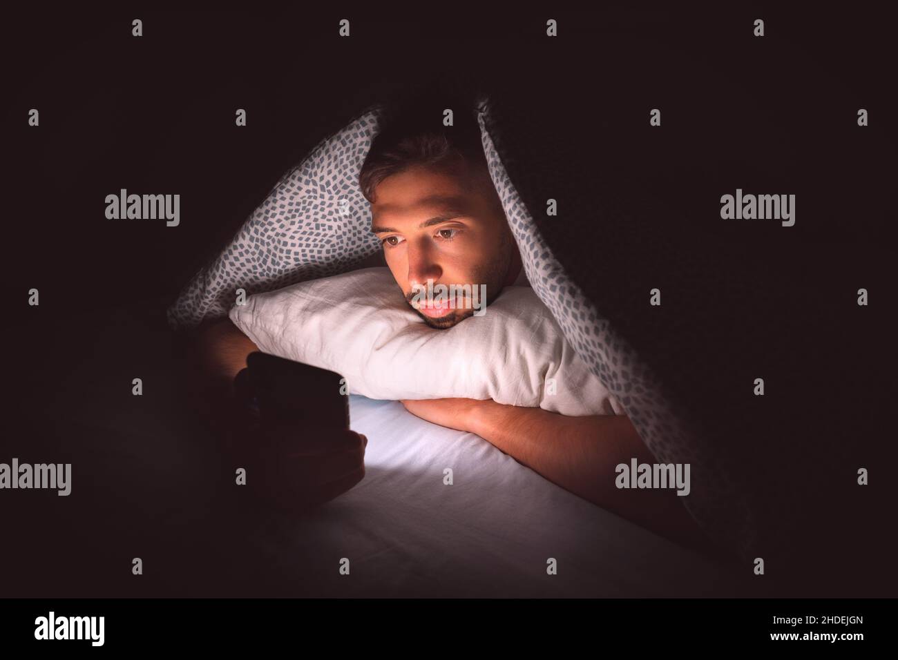 Giovane uomo sdraiato a letto sotto una coperta e con smartphone di notte. Dipendenza dai social media e comunicazione Foto Stock