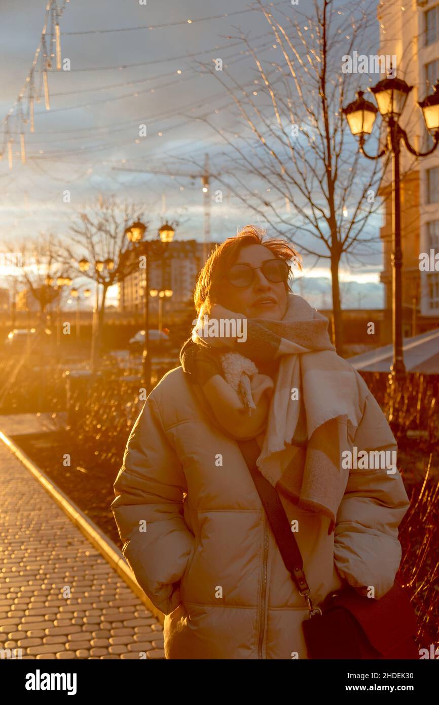 Bella donna boomer bambino di mezza età in occhiali da sole e abiti casual elegante per la strada. Sullo sfondo del tramonto. Foto Stock