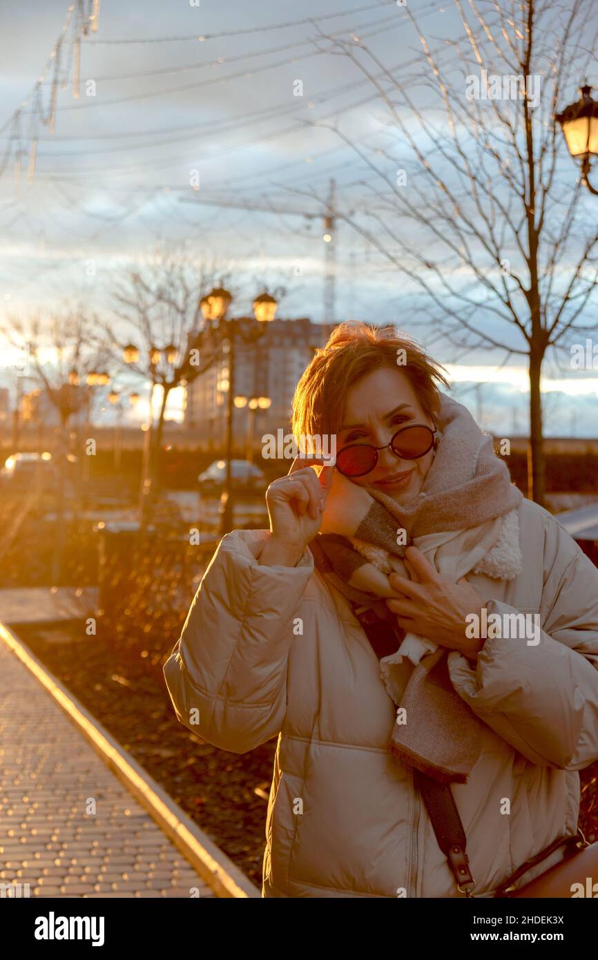 Bella donna boomer bambino di mezza età in occhiali da sole e abiti casual elegante per la strada. Sullo sfondo del tramonto. Foto Stock