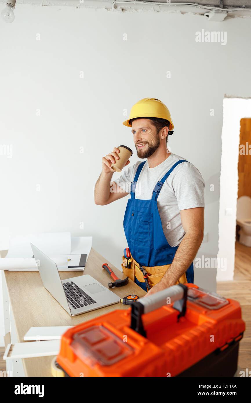 Un uomo rilassato in uniforme e un cappello che beve caffè durante una pausa mentre si svolgono lavori di ristrutturazione al chiuso Foto Stock