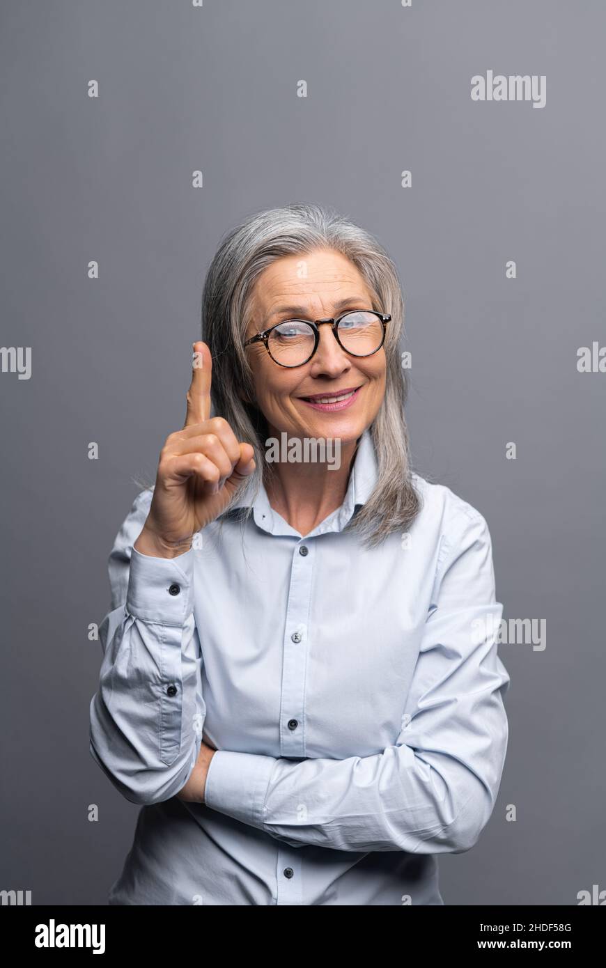 Colpo verticale del sorridente maturo grigio-capelli donna di affari isolato su sfondo grigio punti dita su. Signora che presta attenzione a qualcosa Foto Stock