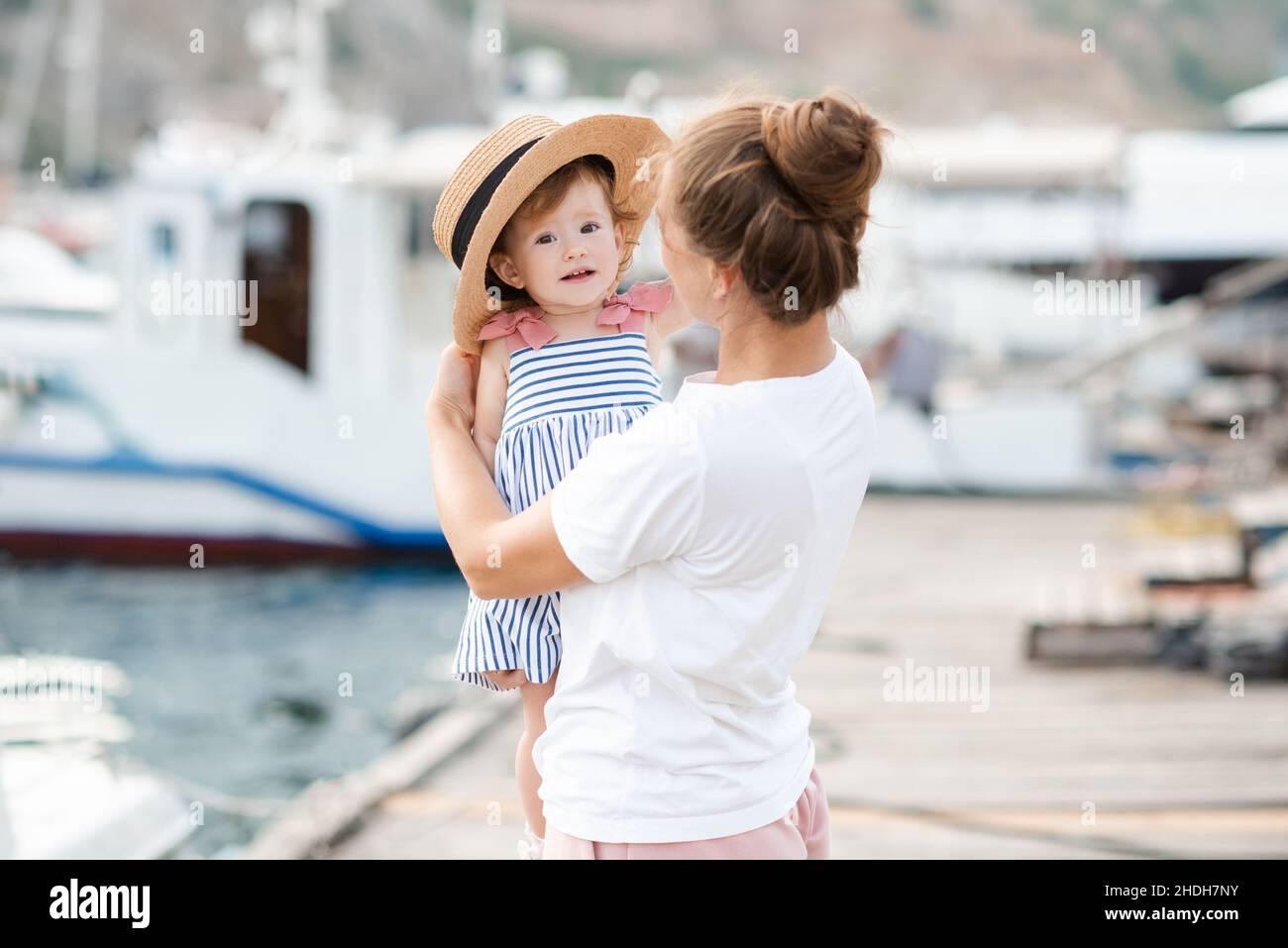 Madre con bambina 1-2 anni con cappello di paglia sul mare e barche a sfondo da vicino. Stile di vita familiare. Stagione estiva. Infanzia. Foto Stock