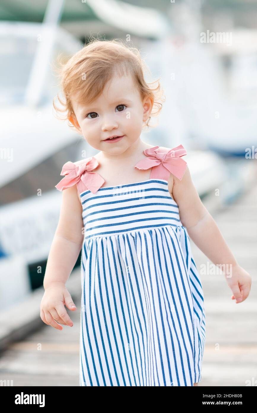 Sorridente bambina di 1-2 anni indossare abito a strisce e tenere camminare sopra il molo di legno sulla riva del mare all'aperto. Stagione estiva. Infanzia. Guardando la fotocamera Foto Stock