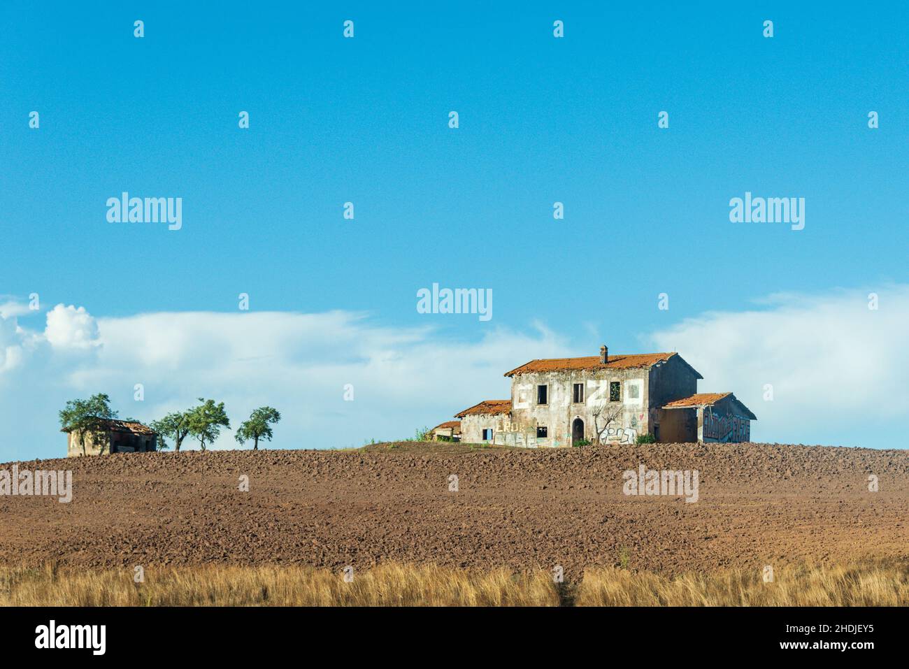 Casa abbandonata sulla collina. Edificio in mattoni ricoperto di piastrelle rosse, finestre e porte rotte. Intorno al campo arato. Ed alcuni alberi verdi Foto Stock
