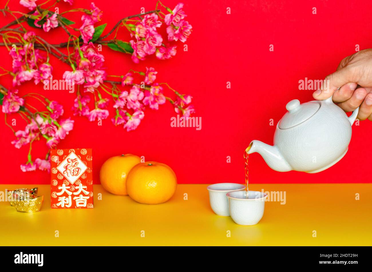 Versare il tè a mano dalla pentola per il concetto cinese di nuovo anno con arance, busta rossa o ang bao (parola significa grande fortuna) e lingotti su gl giallo e rosso Foto Stock