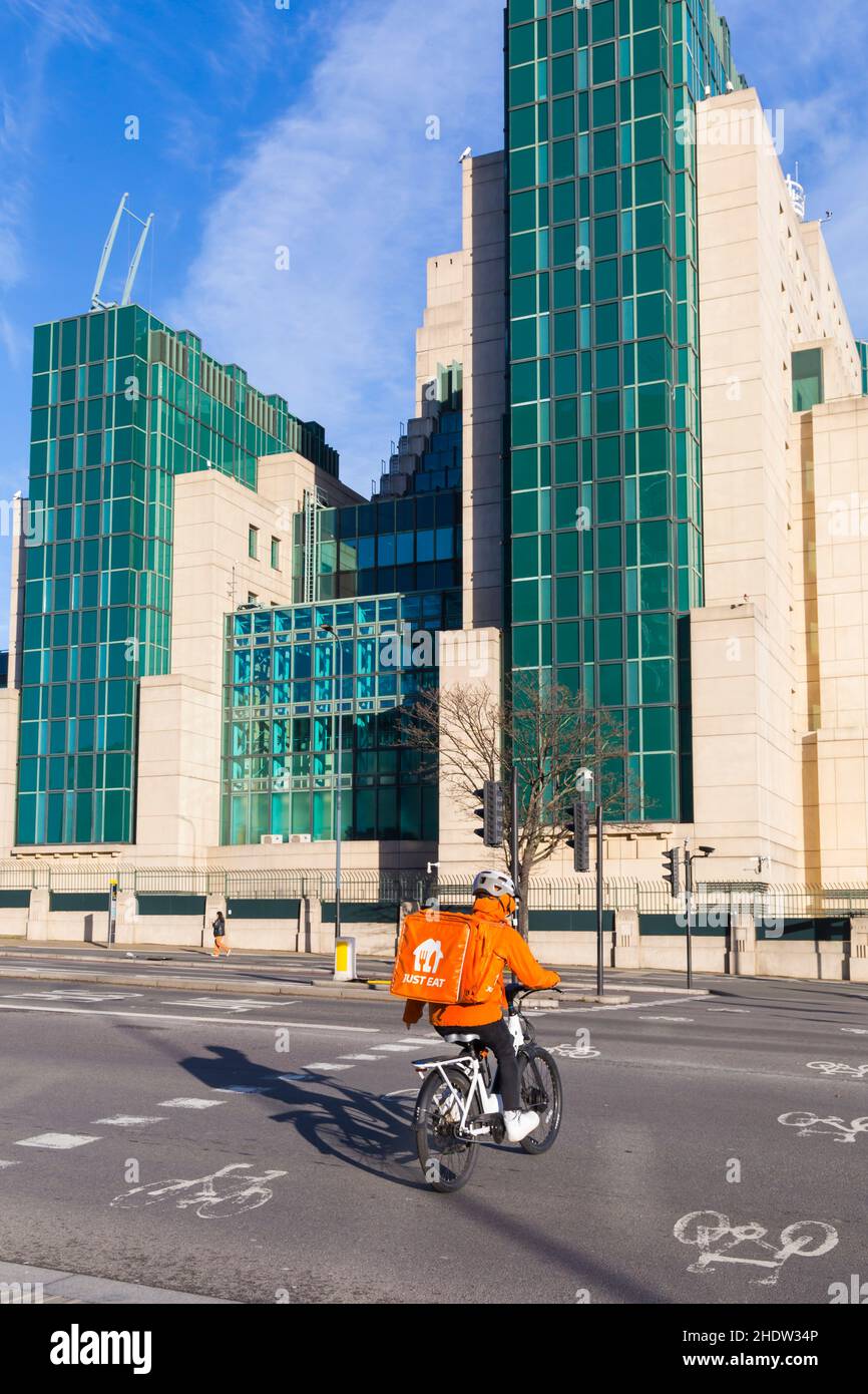 Un pilota di consegna Just Eat su una bicicletta attraversa una pista ciclabile a un bivio, con MI6 edificio sul lato opposto a Vauxhall, Londra UK nel mese di dicembre Foto Stock