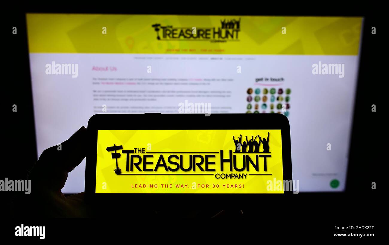 Persona che tiene uno smartphone con il logo della Treasure Hunt Company (Brighton) sullo schermo di fronte al sito Web. Mettere a fuoco sul display del telefono. Foto Stock