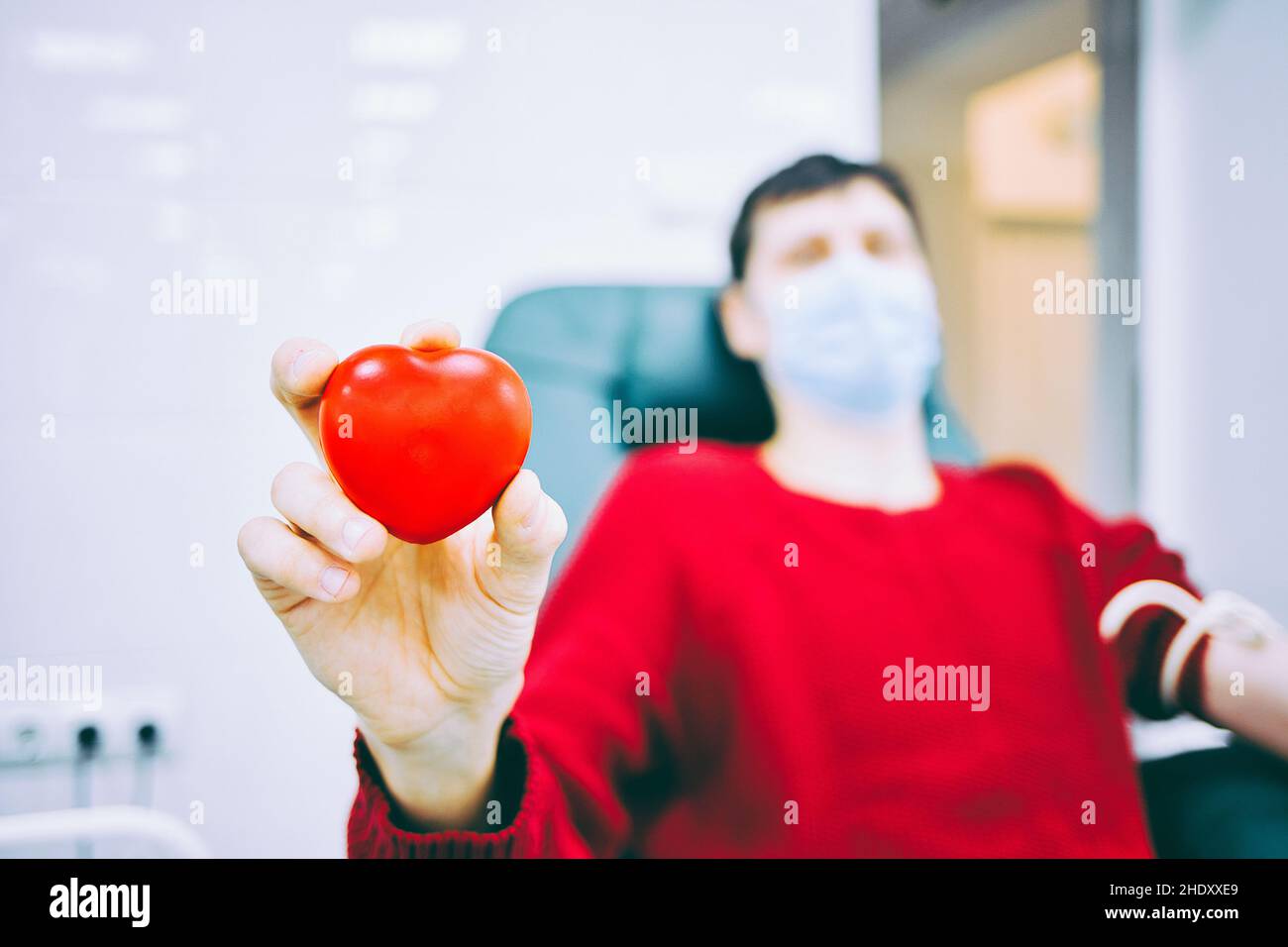 L'uomo si siede in sedia e dona sangue in clinica medica. Tema della donazione. Il donatore tiene in mano il cuore di plastica. Simbolo del donatore. Assistenza gratuita a chi ne ha bisogno. Foto Stock